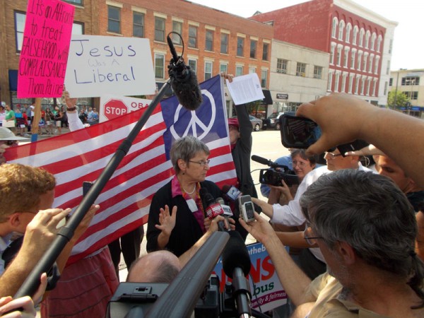 Nuns on the Bus supporters hold up a peace flag behind Sr Simone, to block the view of the "Stop the HHS Mandate"/"Stand Up For Religious Freedom" signs. Here, the "stop sign" peeks out from behind the flag.